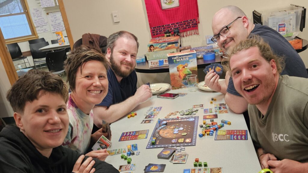 Common Flame members and friends sit around a table playing a board game during our community board game night and potluck
