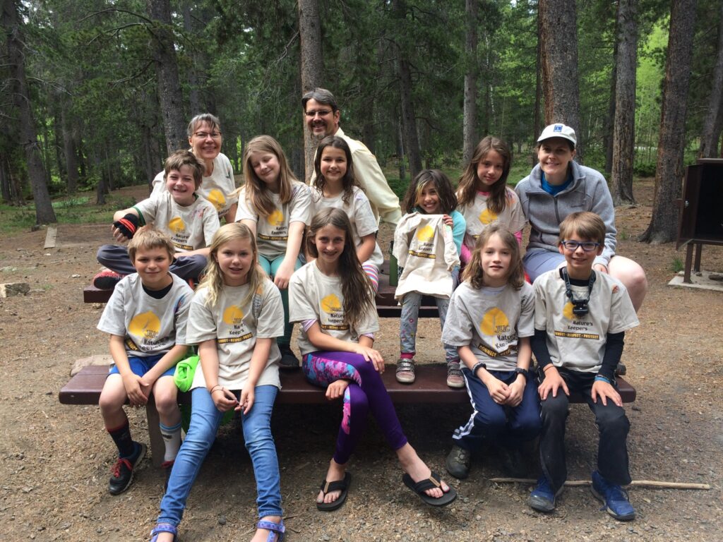 A group of kids attending a camping trip pose for a photo while sitting on a picnic table 